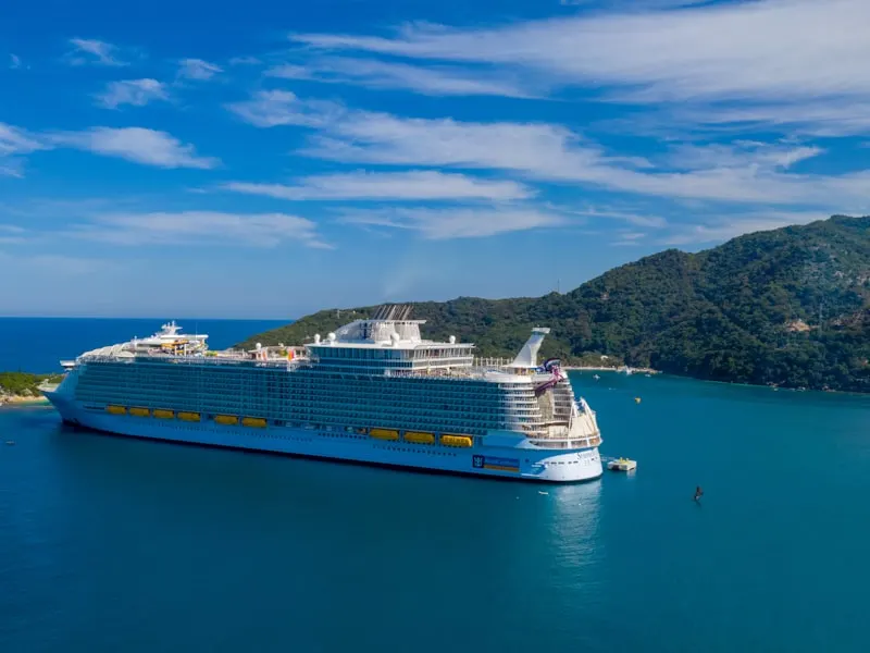 Royal Caribbean ship by a forested bay, with line branding visible